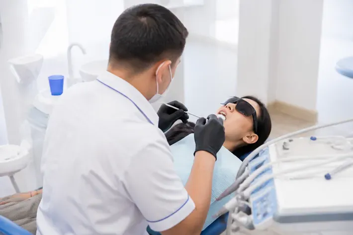Chairside view: dentist at work, shot over the assistant's shoulder — illustration for the section "What types of lasers are used in dentistry and how do they differ"