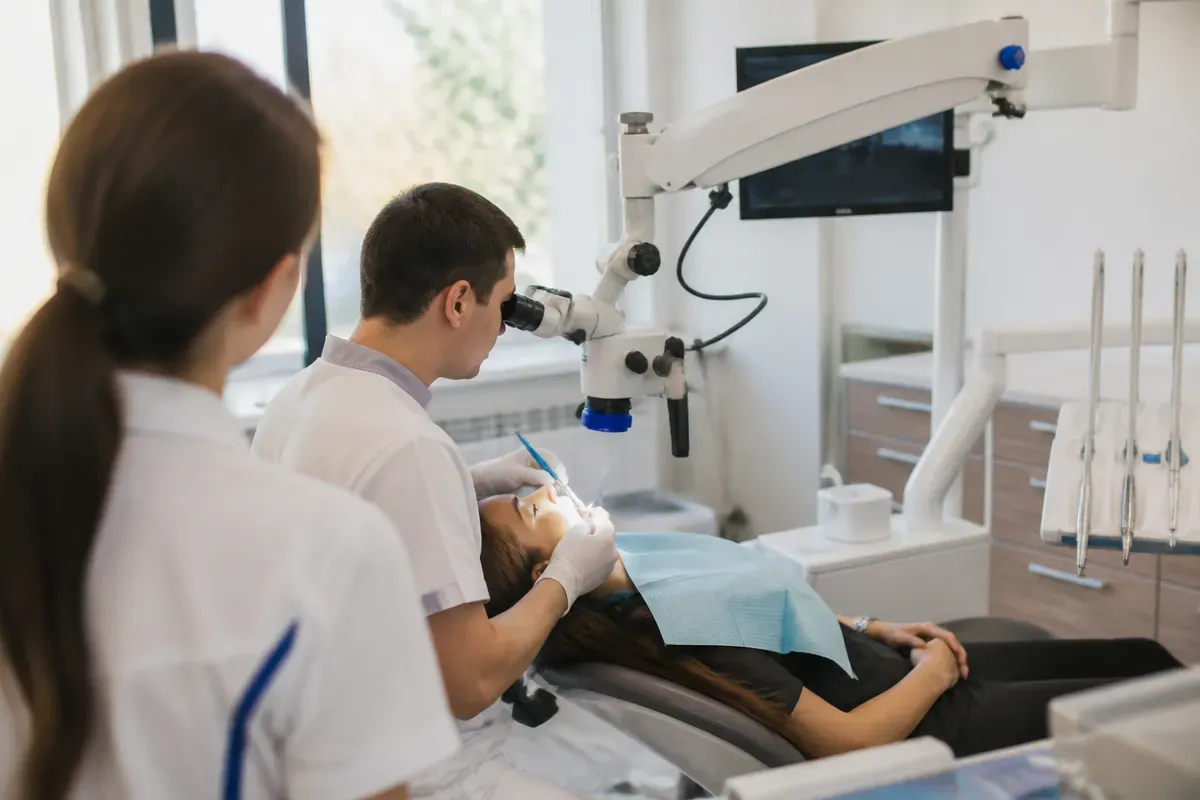 Орындық жанындағы қабылдау: жұмыс үстіндегі дәрігер, көмекшінің иығынан түсірілген сурет — «Пульпит кезіндегі асқынулар» бөліміне иллюстрация