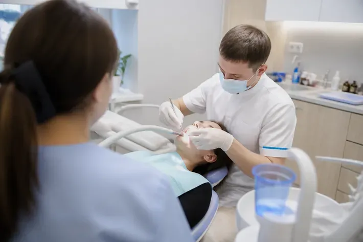 Орындық жанындағы қабылдау: жұмыс үстіндегі дәрігер, көмекшінің иығынан түсірілген сурет — «Стоматологияда қандай лазерлер қолданылады» бөліміне иллюстрация