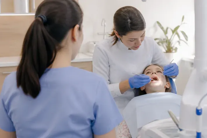 Chairside appointment: dentist at work, over-the-shoulder shot past the assistant — illustration for the section "Crowns for Baby Teeth"